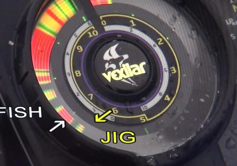 A close-up of a Vexilar fish finder display shows colored bars indicating sonar readings, essential for fishing. The words "FISH" and "JIG" with arrows point to different bars on the wet dial, dotted with visible water droplets.