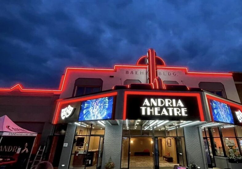 The Andria Theatre building at night, outlined with red neon lights, glows under a dramatic dark blue sky. The Andria Theatre’s entrance is lit up, with digital screens displaying graphics on both sides of the theater sign.