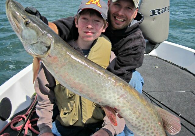 Two people sitting on a boat, smiling and holding a very large fish—likely a muskie—caught using mid-summer fishing tactics, with a lake and trees in the background under a partly cloudy sky.