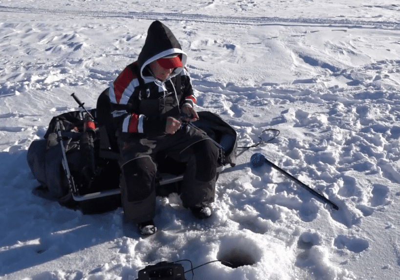 A person in winter clothing sits on a sled, panfishing on a snowy frozen lake. They hold fishing gear near a hole in the ice, with an auger and other equipment nearby.