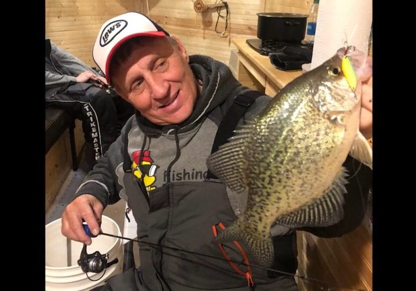 A smiling man, mike frisch, in fishing gear holds up a large speckled fish inside a wooden cabin, showing it to the camera. He has a fishing rod in one hand and wears a white cap with logos.