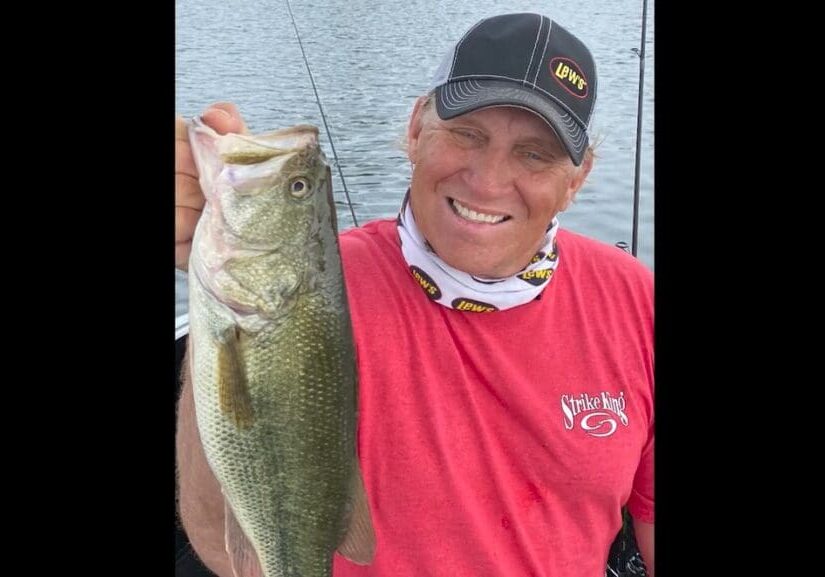 A smiling man in a red shirt and black cap holds up a large fish he caught while fishing, standing on a boat with water in the background.