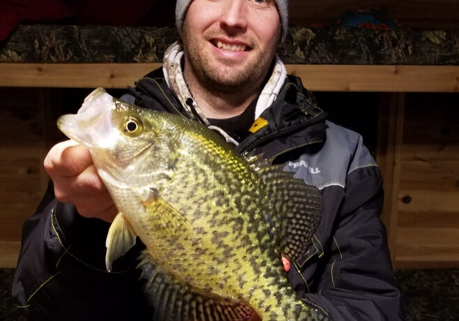 A smiling man wearing a gray beanie and a dark jacket proudly holds up his fishing catch—a large yellow and black speckled fish—indoors, with wooden bunk beds in the background.