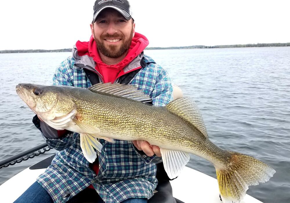 A smiling man wearing a plaid jacket and red hoodie sits on a boat, holding a large walleye fish during the fishing opener, with a lake and cloudy sky in the background.