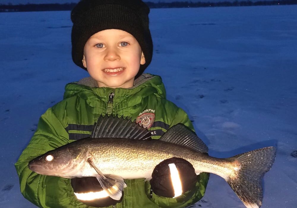 A young child in a green jacket and black hat smiles while holding a large walleye with both hands, standing on a snowy, frozen lake near Alexandria at dusk.