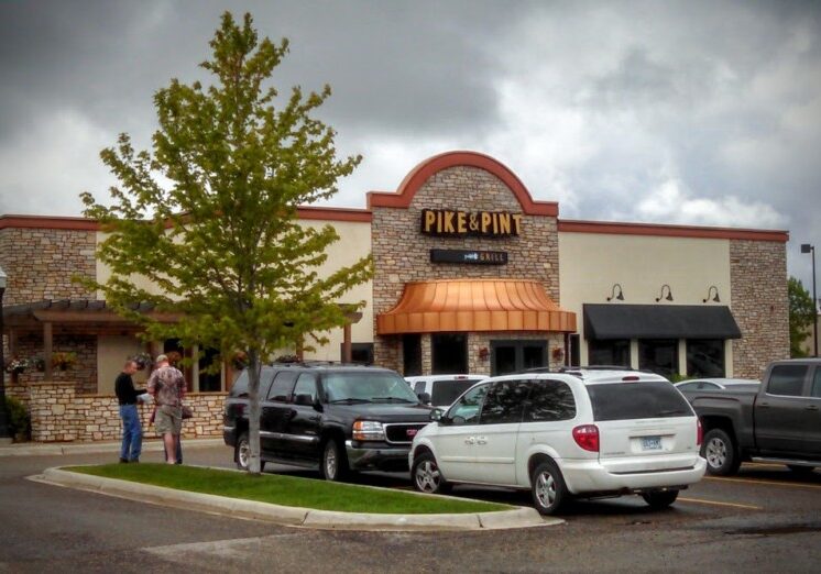 A bar and grill restaurant named "Pike & Pint" features a stone and stucco exterior, several parked cars in front, and three people standing near the entrance on a cloudy day.