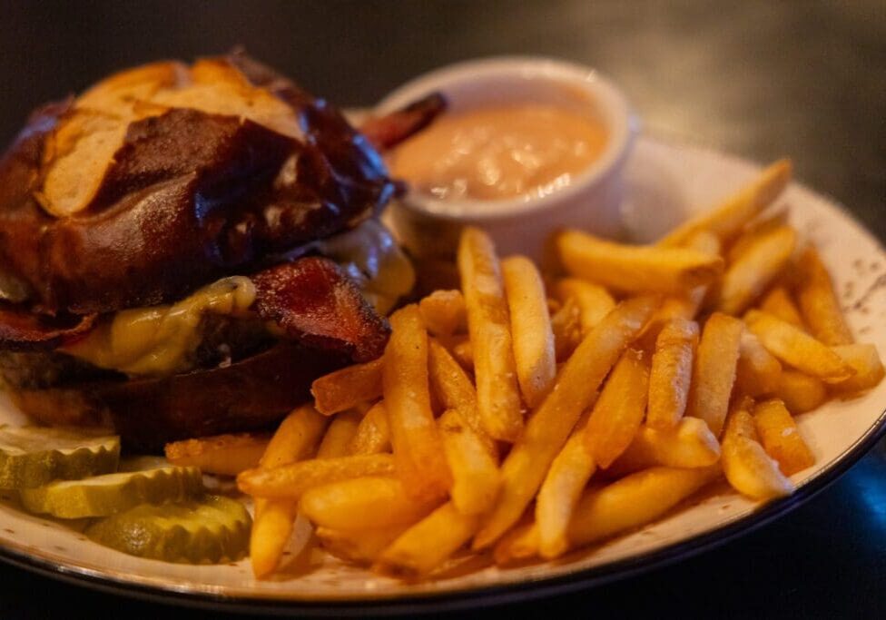 A plate with a cheeseburger on a pretzel bun, crispy French fries, pickle slices, and a small cup of dipping sauce—classic comfort food you’ll find at top Alexandria MN restaurants.