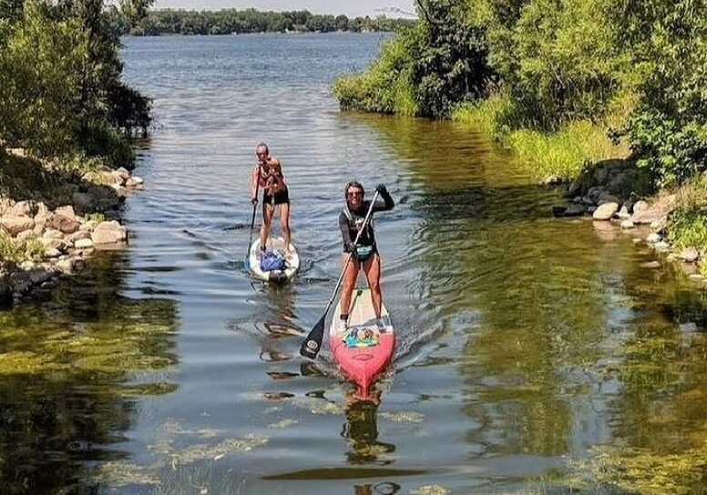 Two people paddleboarding on a calm, narrow waterway surrounded by green trees and grass, heading toward an open lake under a blue sky with scattered clouds during the Big Ole Paddlefest event.