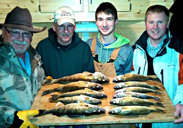 Four men indoors smile at the camera while holding a wooden board with ten freshly caught fish arranged in two rows—a great catch featured in this January fishing report. The background showcases wooden cabinetry, highlighting an Alexandria fishing success.
