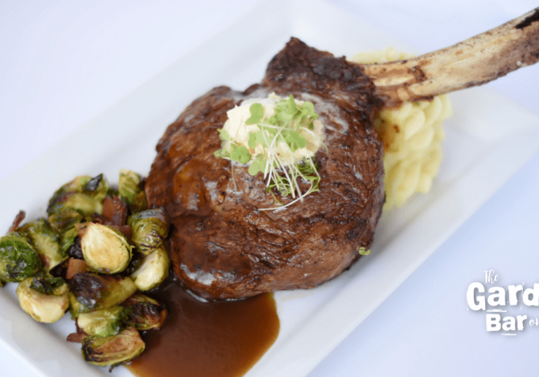 A grilled tomahawk steak topped with microgreens and butter, served with mashed potatoes, roasted Brussels sprouts, and brown sauce on a white plate—classic flavors from one of the finest restaurants in Alexandria MN. “The Garden Bar on 6th” logo appears in the corner.