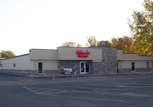 A single-story building with tan walls and stone accents, displaying a red neon sign reading “Route” above the entrance. The mostly empty parking lot sits in front of Ron's Warehouse, framed by trees with autumn foliage in the background.