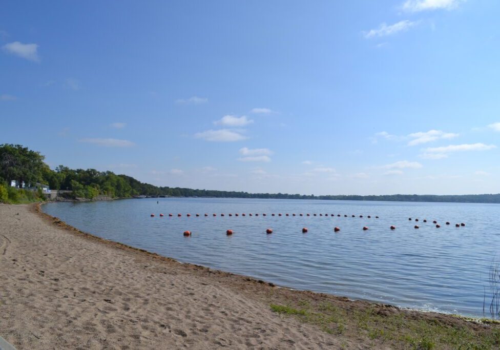A sandy beach curves along calm lake waters at Rotary Beach Alexandria MN, with a line of red buoys floating under a clear blue sky and green trees surrounding Lake Le Homme Dieu.