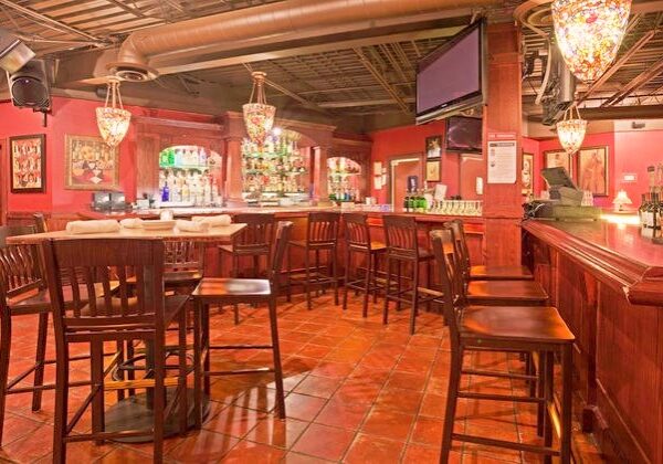 A cozy bar, Willy T's, with high wooden chairs and tables, warm lighting from hanging lamps, shelves of liquor bottles behind the bar, red walls, and framed artwork creates an inviting atmosphere.