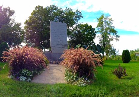 A stone monument stands in Runestone City Park, surrounded by ornamental grasses, small flowering plants, and trees beneath a partly cloudy sky.