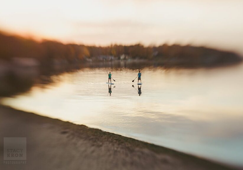 Two people stand on paddleboards in calm water at sunset, each holding a paddle, as soft focus photography blurs the surroundings and reflects the warm sky and shoreline in the background.