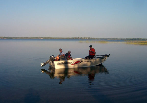 Three people sit in a small motorboat labeled "WARRIOR BOAT" on calm, reflective water under a clear blue sky, enjoying a scenic view of the distant shoreline and trees—perfect for those visiting the nearby RV campground.