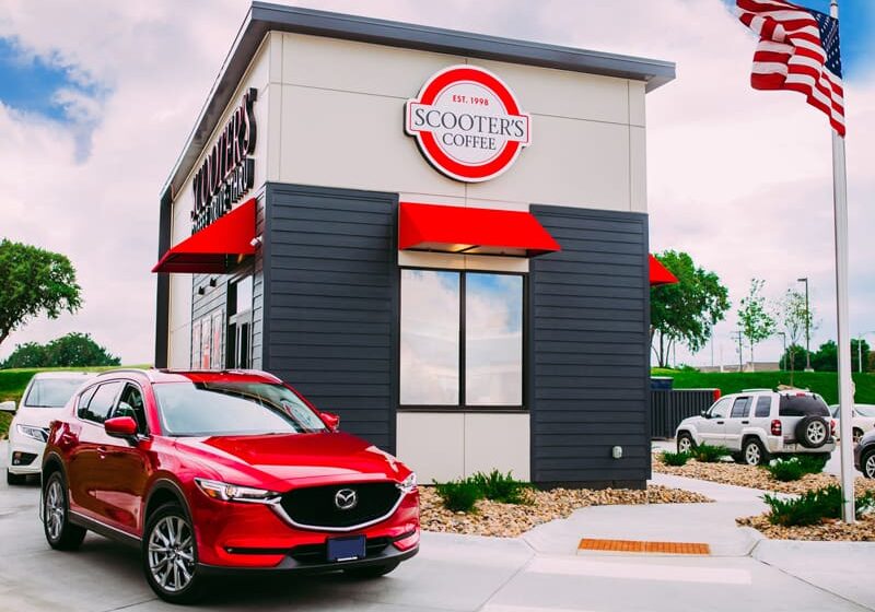 A red SUV waits at the drive-thru of Scooter’s Coffee, a modern café with bold red accents. The American flag waves nearby, while greenery surrounds Scooter's Coffee under a partly cloudy sky.
