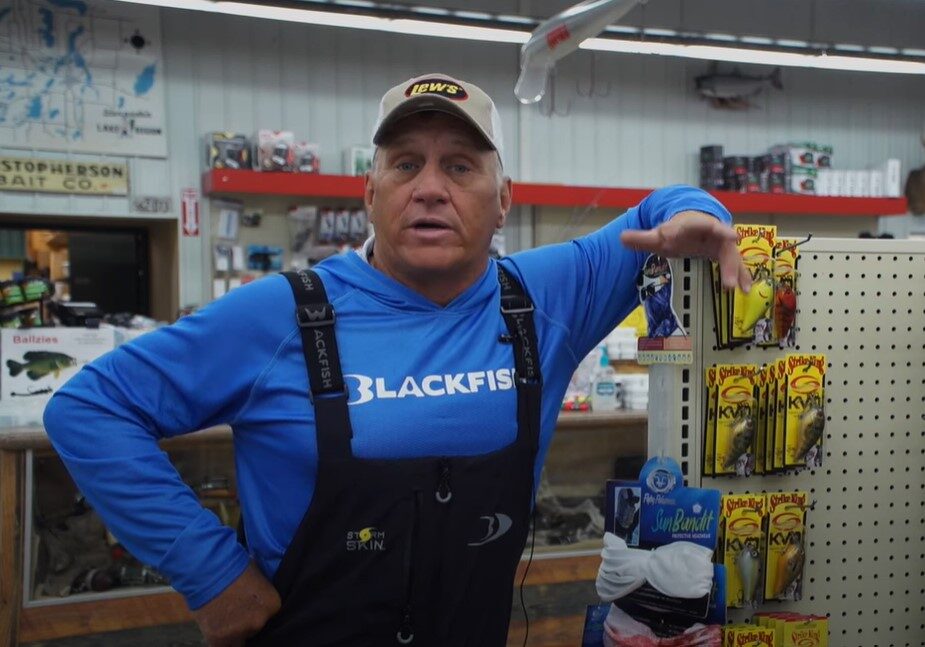 A man in a blue "Blackfish" shirt and overalls leans on a store display stocked with bass fishing lures. He wears a white cap and stands in a fishing supply shop filled with gear, signs, and mounted fish on the walls.