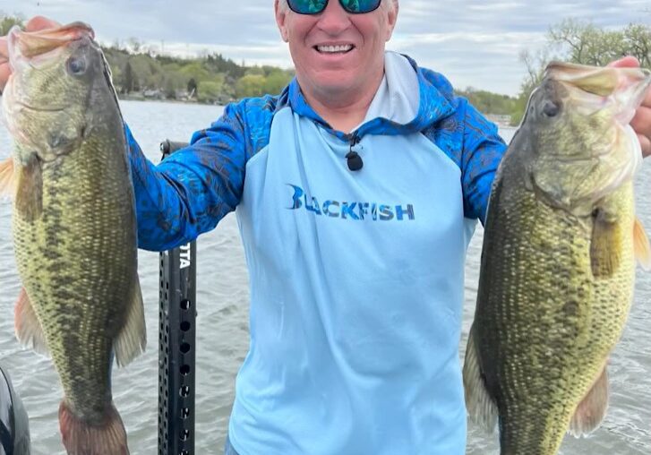 A smiling man in a blue fishing shirt and cap holds up two large bass fish, showcasing his season highlights while standing on a boat in a lake. Trees and water are visible in the background.