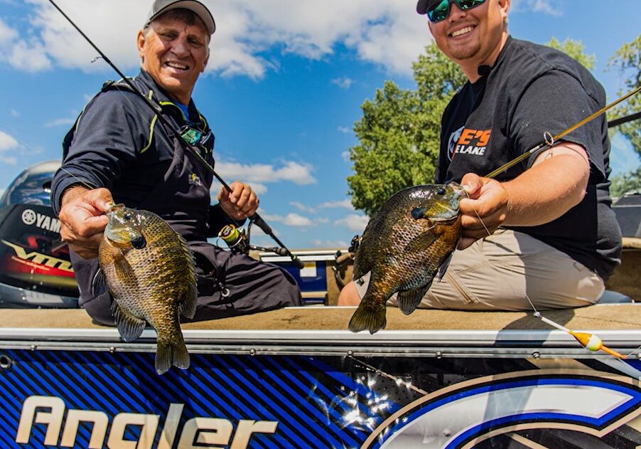 Two smiling men sit in a boat labeled “Angler v1850,” holding up freshly caught fish. Enjoying multi-species fishing in the Alexandria area, they wear sunglasses and caps with blue sky and trees in the background.