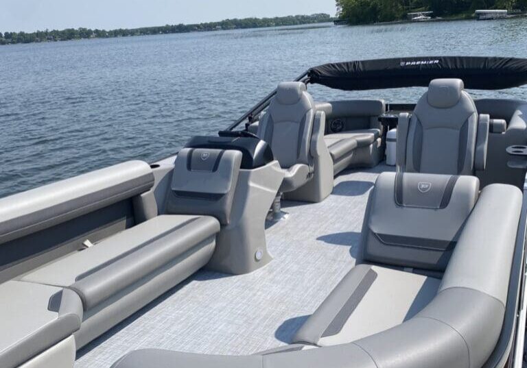 A spacious pontoon boat with gray and beige cushioned seating floats on a calm lake near Betsy Ross Resort, surrounded by trees and a distant shoreline under a clear sky.