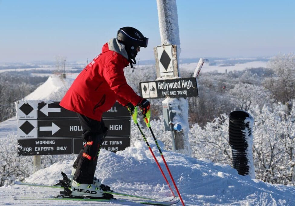A skier in a red jacket and black helmet stands outdoors at the top of a snowy slope near signs that read “Hollywood High” and “For Experts Only,” with frosty trees and a blue sky in the background.