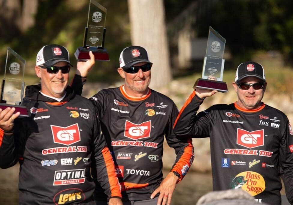 Three men wearing matching black and red General Tire fishing jerseys and hats smile while holding up clear glass trophies outdoors, with trees and sunlight in the background after a successful day of bass fishing.