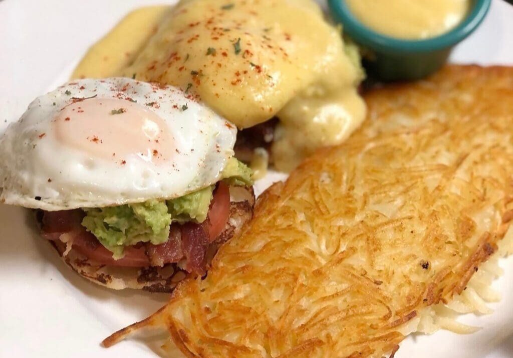 A delicious RESTAURANT WEEK breakfast plate featuring eggs Benedict with hollandaise, a fried egg with avocado and bacon on an English muffin, crispy hash browns, and a small cup of yellow sauce—perfect for breakfasts lovers.