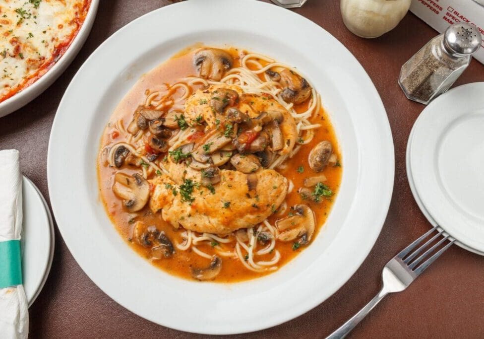 A plate of pasta topped with chicken, mushrooms, and a tomato-based sauce, garnished with herbs. Capturing the essence of Tuscany, the scene evokes an authentic Italian restaurant experience with parmesan cheese and classic table settings nearby.