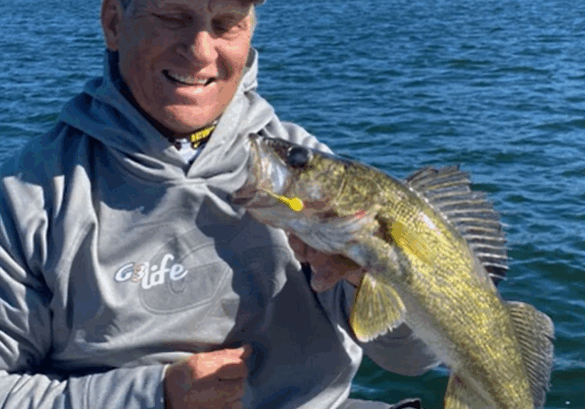 A smiling person in a gray hoodie and baseball cap holds up a large yellow-green fish on a boat, enjoying fishing the Midwest on one of Alexandria MN’s picturesque lakes, with autumn foliage and a blue lake in the background.