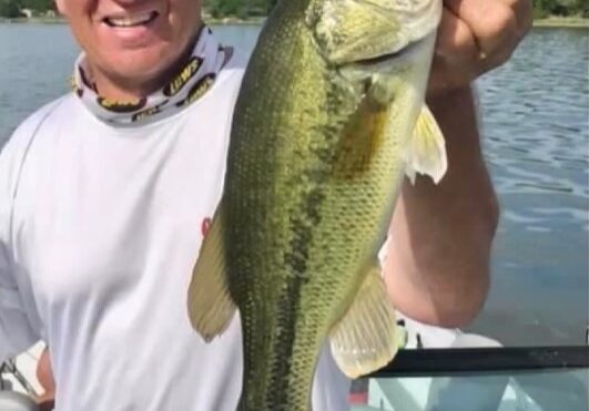 A man wearing a cap and white shirt smiles while holding a large bass fish in one hand, standing on a boat with water and trees in the background—enjoying fishing weed lines during the dog days of summer.