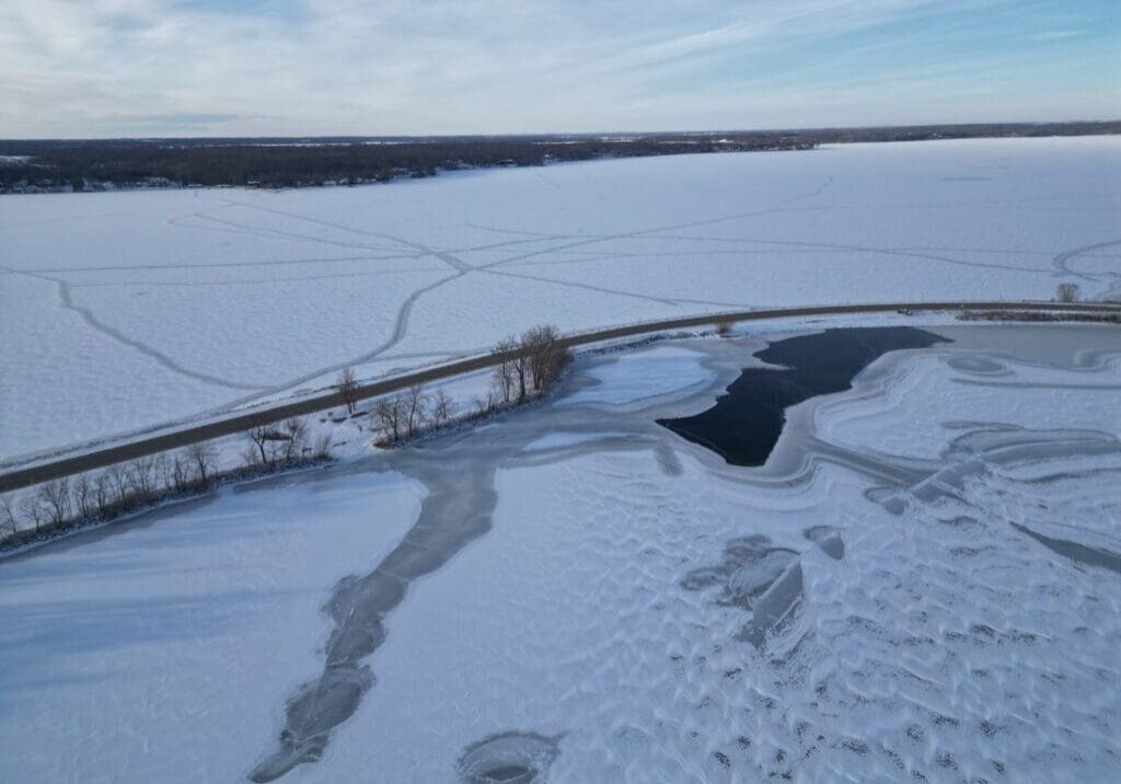 A frozen lake in the Alexandria Lakes Area features snow-covered ice, intersected by a road; winding tire or walking tracks cross the snowy surface, with open water near the shore and a partly cloudy blue sky above.