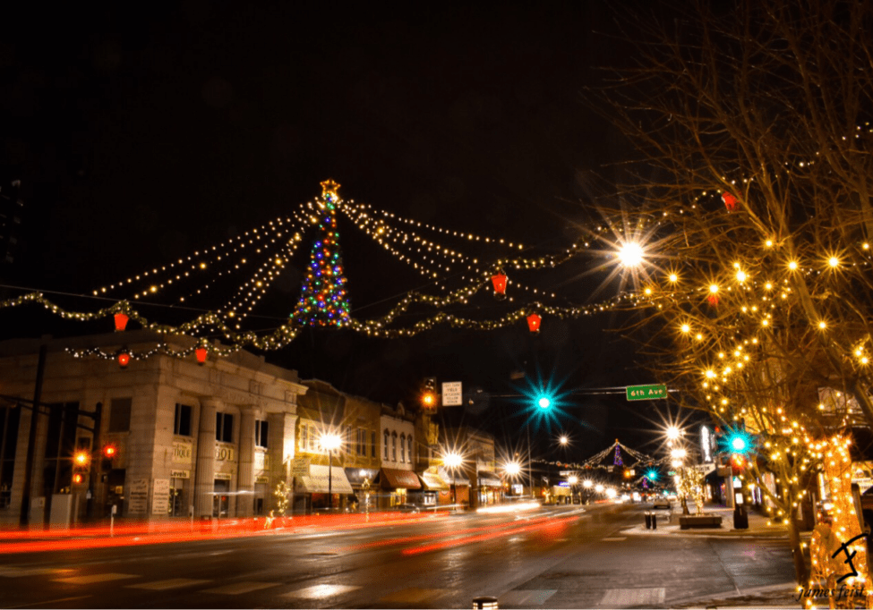 A downtown street at night is decorated with Christmas lights, including a lit tree and string lights above, as storefronts glow for holidays shopping and car light trails line the road—reminding everyone to shop safe this season.
