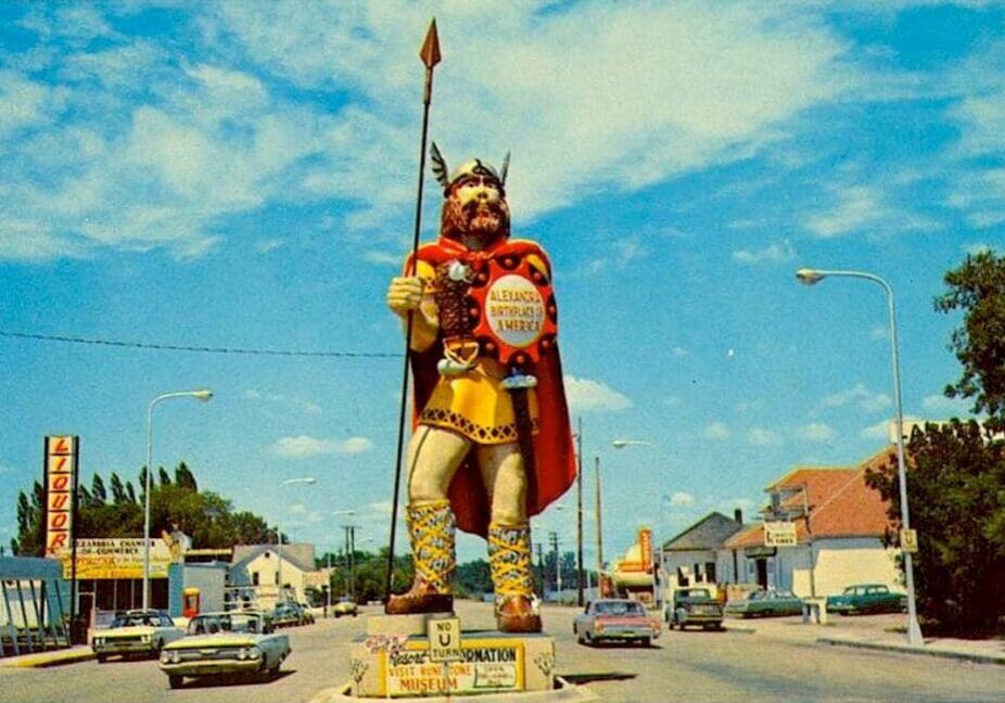 A large, colorful statue of a Viking warrior—known as "Big Ole" through the years—stands on a street median, surrounded by vintage cars, streetlights, and buildings under a blue sky with scattered clouds.