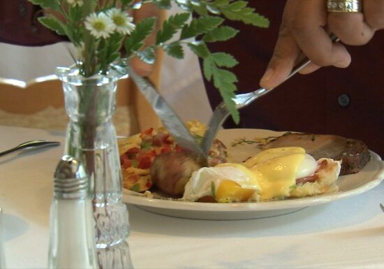 At a charming Bed & Breakfast, a person cuts into eggs benedict with home fries and roast meat, set on a white tablecloth alongside a glass of water, flower vase, and salt and pepper shakers.