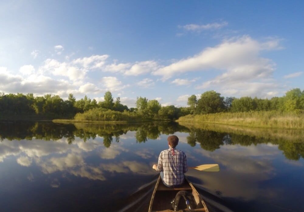 A person in a plaid shirt paddles a canoe on a calm lake in the Alexandria Lakes Area, surrounded by trees and greenery, with blue sky and clouds mirrored on the water—one of the 20 must-do summer activities in this scenic region.