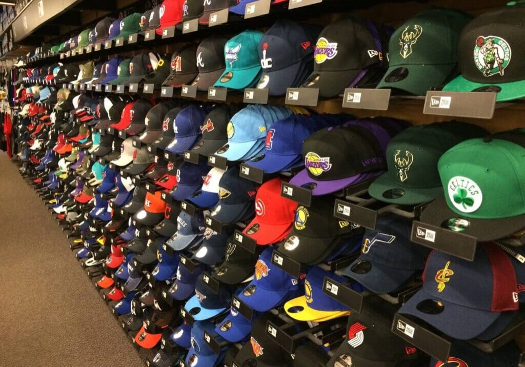 Rows of colorful baseball caps with various NBA team logos are neatly displayed on shelves in the Cowing Robards store, covering several teams and colors, creating a vibrant and organized layout.