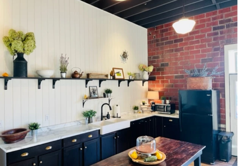 A modern kitchen at The Creamery & Corner Garage features dark cabinets, white countertops, exposed brick walls, open shelves with plants and kitchenware, black appliances, and a wooden table with a cake stand and lemons.