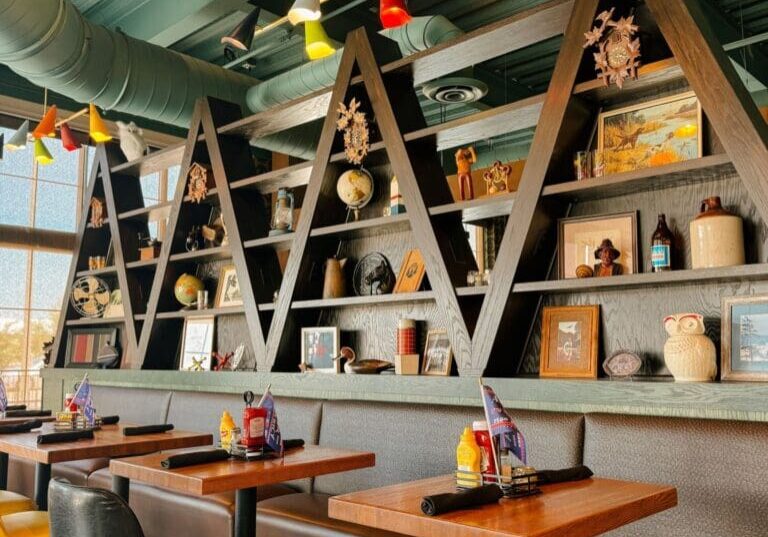 A modern restaurant interior with wooden tables, yellow chairs, and triangular shelves holding various decorative items like vases, books, and figurines. Bright pendant lights hang from the ceiling, while large windows offer views of tall Longtrees just outside.