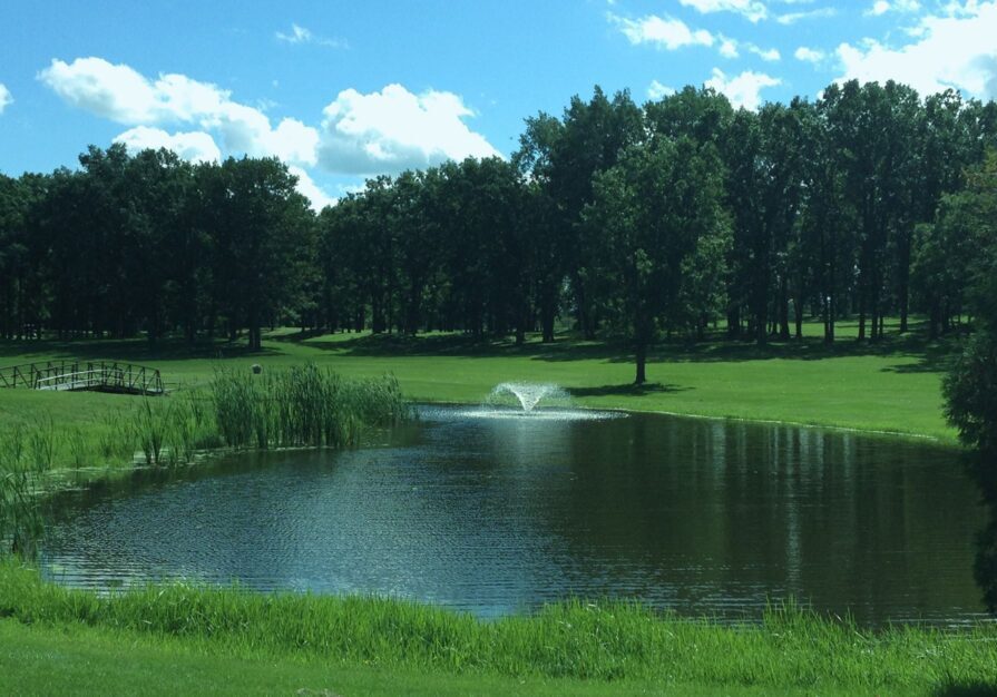 A peaceful pond with a small fountain sits in a grassy park near Double Eagle Golf, surrounded by trees. A wooden footbridge crosses a narrow section of the pond under a sunny blue sky with scattered white clouds.