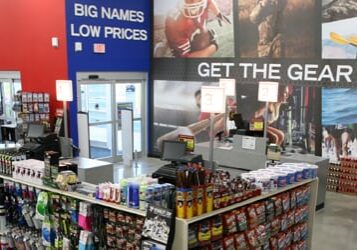 Wide view of a Dunham's Sports store interior, featuring shelves stocked with gear and accessories, colorful kayaks on the left wall, and large wall graphics promoting outdoor activities and top sports brands.