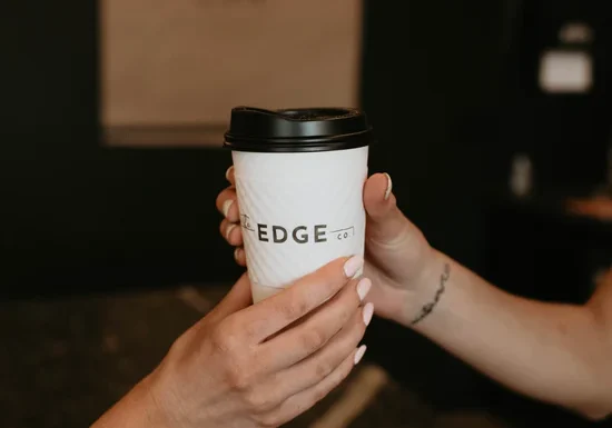 Two hands exchange a white takeaway coffee cup with a black lid labeled "EDGE co." from The Edge Co. against a dark, blurred background.