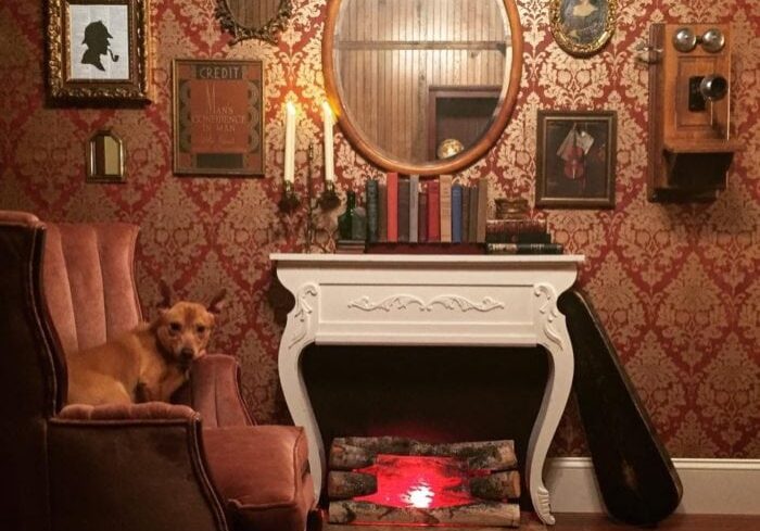 A cozy vintage Escape Room Alexandria with ornate red wallpaper, a faux fireplace, antique decor, framed pictures, books, and a brown dog sitting on a velvet armchair. Logs with red lights imitate a fire in the fireplace.