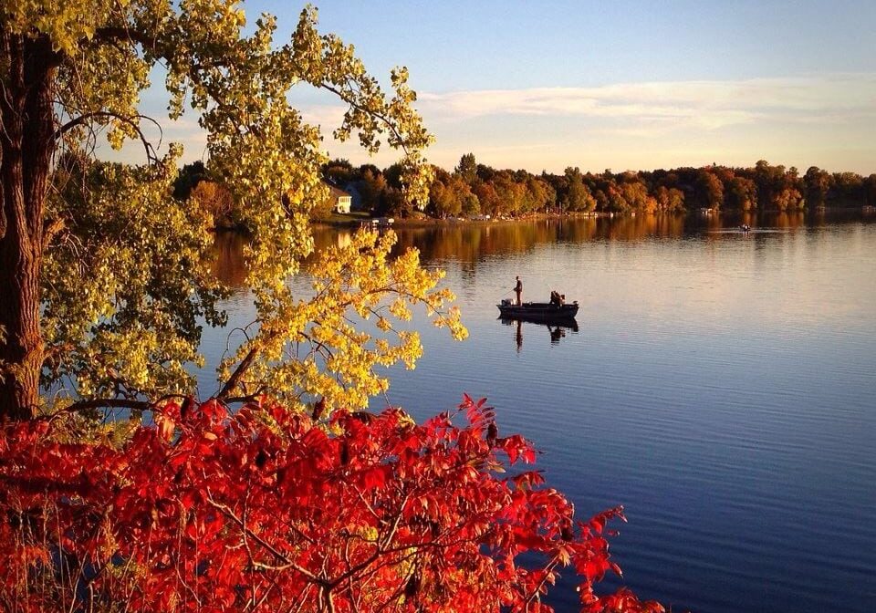 A calm lake reflects autumn trees with red, yellow, and green leaves. Two people fish in a small boat under a clear blue sky—just one of the 15 ways to explore Alexandria MN in October.