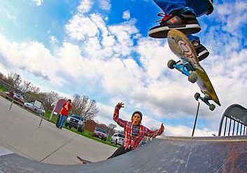 A skateboarder in a striped shirt performs a trick in mid-air above a ramp at Dean Melton Fillmore Park, while another person in a red plaid shirt reaches up from below. The scene unfolds outdoors under a partly cloudy sky.