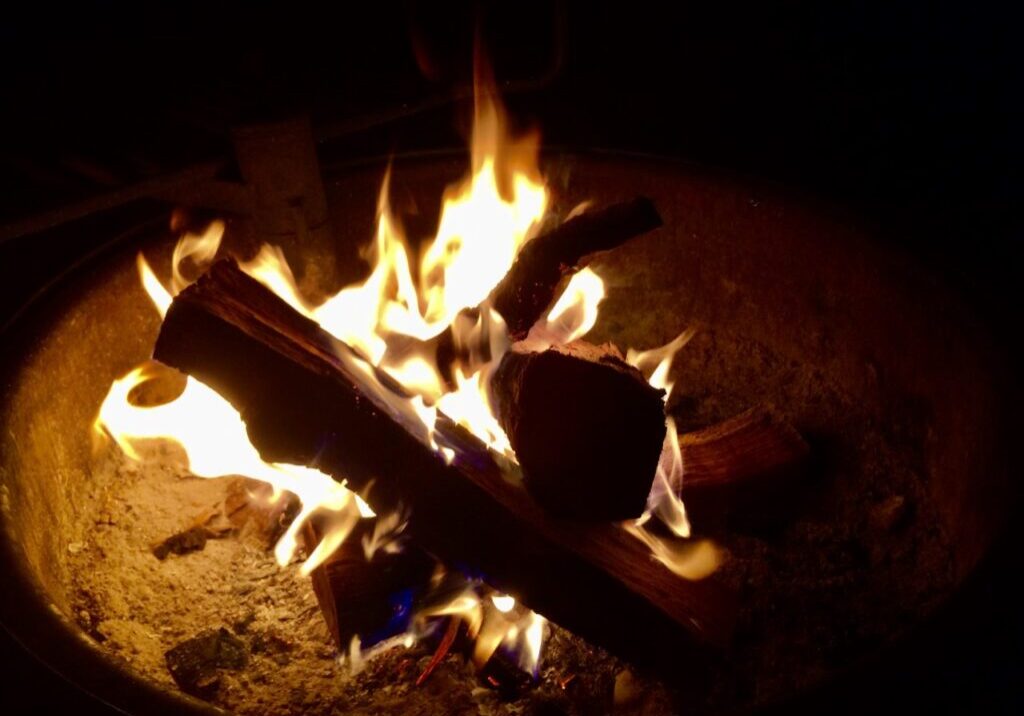 A close-up of a campfire at night in the Lakes Area, with glowing orange flames rising from burning logs arranged in a fire pit, surrounded by ash and embers—perfect for socially distant summer activities.