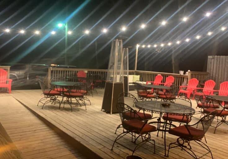 An outdoor wooden deck at night with string lights overhead at Fired Up Bar & Grill, featuring black metal tables and chairs with red cushions, red Adirondack chairs along the railing, and a well-lit, inviting atmosphere.