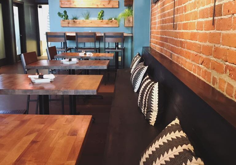 A cozy Garden Bar on 6th interior with wooden tables, patterned black and white pillows on a bench, a brick wall, blue accent wall, and shelves with potted plants. Soft lighting creates a warm, inviting atmosphere.