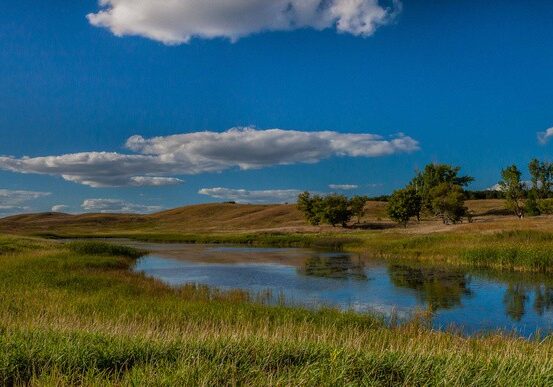 A calm river winds through grassy plains under a bright blue sky with scattered white clouds in Glacial Lakes State Park. Gentle hills and a few trees are visible in the background on a clear, sunny day.
