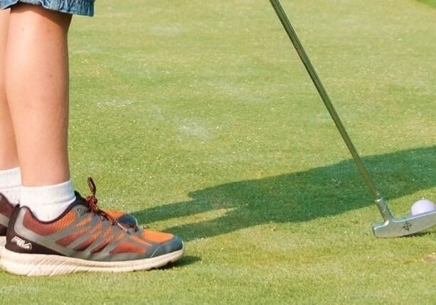 A person in sneakers and shorts stands on a green, preparing to hit a golf ball with a putter—a classic scene for anyone getting started in golf. Only their legs and the golf club are visible.